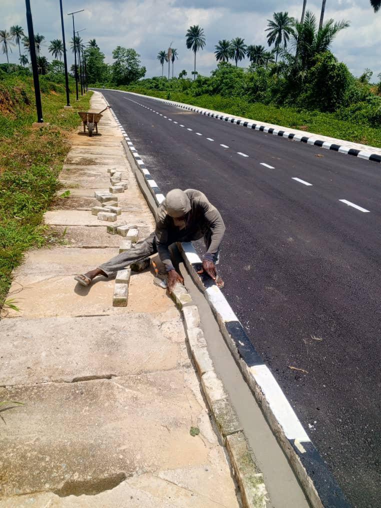 Worker finishing the painted kerb edge by hand beside the completed road, with lane markings running into the distance and a wheelbarrow on the sidewalk